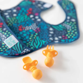 A Bumkins SuperBib® 3 Pack: All Together Now lays out of focus on a white background, with orange silicone utensils in the foreground.