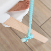 Load image into Gallery viewer, Close-up of a baby's foot in a high chair, with a spoon hanging from a Bumkins Silicone Accessory Tether 2-Pack: Blue above the wood floor.
