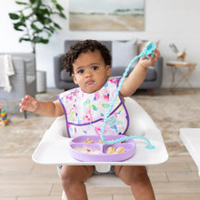 Load image into Gallery viewer, A toddler in a high chair wears a colorful bib, holding a spoon attached to the tray with a Bumkins Silicone Accessory Tether 2-Pack: Blue.

