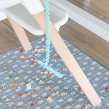 Load image into Gallery viewer, A spoon secured by a Bumkins Silicone Accessory Tether 2-Pack: Blue dangles from a high chair, as food rests on a patterned mat below.
