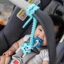 Load image into Gallery viewer, A smiling child chews a Bumkins Silicone Teething Charms: Blue attached to a blue silicone tether in a car seat.
