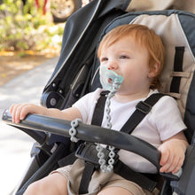 Load image into Gallery viewer, A baby in a stroller outdoors using a pacifier attached to a Bumkins Silicone Accessory Tether 2-Pack: Gray, with greenery behind.
