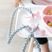 Load image into Gallery viewer, Close-up of pink toddler utensils attached to Bumkins Silicone Accessory Tether 2-Pack: Gray on a high chair tray.
