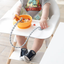 Load image into Gallery viewer, A baby in a bib and sneakers eats from a bowl in a high chair, with a spoon attached to a Bumkins Silicone Accessory Tether 2-Pack: Gray.

