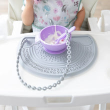 Load image into Gallery viewer, A toddler enjoys yogurt from a purple bowl on a gray mat with a Bumkins Silicone Accessory Tether 2-Pack: Gray attached to a spoon.

