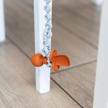 Load image into Gallery viewer, Close-up of an orange toddler spoon hanging from a Bumkins Silicone Accessory Tether 2-Pack: Gray next to a white chair leg on a wood floor.
