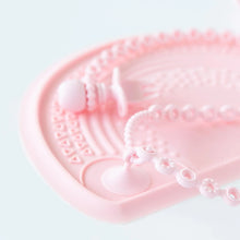 Load image into Gallery viewer, Close-up of a Bumkins Silicone Accessory Tether 2-Pack: Pink attached to a pink placemat on a white background, holding a toddler utensil.
