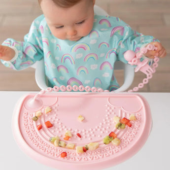 A baby in a rainbow bib eats fruit on a placemat, holding a pink toddler fork attached to a Bumkins Silicone Accessory Tether 2-Pack: Pink.