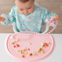 Load image into Gallery viewer, A baby in a rainbow bib eats fruit on a placemat, holding a pink toddler fork attached to a Bumkins Silicone Accessory Tether 2-Pack: Pink.
