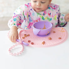 Load image into Gallery viewer, A baby in a bib eats from a purple bowl on a pink mat, holding a purple spoon attached to a Bumkins Silicone Accessory Tether 2-Pack: Pink.
