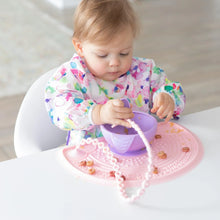 Load image into Gallery viewer, A toddler in a floral bib eats from a bowl on a pink placemat, with a spoon attached to a Bumkins Silicone Accessory Tether 2-Pack: Pink.
