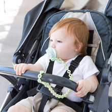 Load image into Gallery viewer, A baby with a pacifier attached to a Bumkins Silicone Accessory Tether 2-Pack: Sage in a stroller, wearing a white shirt and beige shorts.
