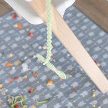 Load image into Gallery viewer, A spoon secured by a Bumkins Silicone Accessory Tether 2-Pack: Sage dangles from a high chair, as food rests on a patterned mat below.
