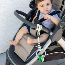 Load image into Gallery viewer, A baby in a stroller joyfully gazes at the camera with a teether on a Bumkins Silicone Accessory Tether 2-Pack: Sage dangling from the side.
