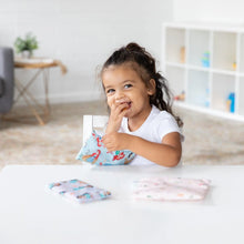 Load image into Gallery viewer, A child beams at a white table with Bumkins Reusable Snack Bag, 3-Pack: Princess Magic, Ariel, and Jasmine in a bright room.
