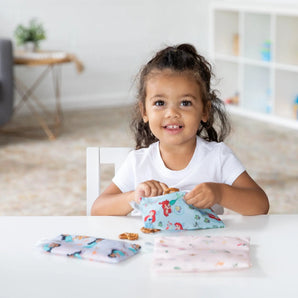 A young girl grins at a table, holding Bumkins Reusable Snack Bag, 3-Pack: Princess Magic, Ariel, and Jasmine in a bright room with shelves.