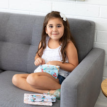 Load image into Gallery viewer, A girl sits on a sofa, holding a Bumkins Reusable Snack Bag, 3-Pack: Princess Magic, Ariel, and Jasmine. Two more bags are beside her.
