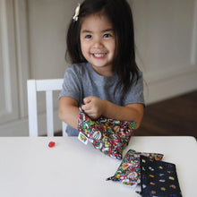 Load image into Gallery viewer, A smiling girl opens a Bumkins Reusable Snack Bag, 3-Pack: Super Mario™ Power Up at a white table.
