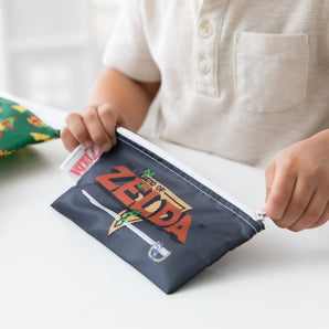 Close-up of a child in white shirt zipping a Bumkins Reusable Snack Bag, 3-Pack: The Legend of Zelda™ at a white table.
