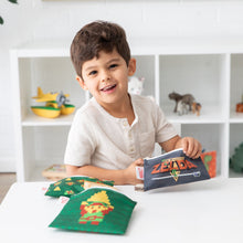 Load image into Gallery viewer, A young boy smiles at a white table with Bumkins Reusable Snack Bag, 3-Pack: The Legend of Zelda™; shelves of toys in the background.
