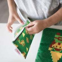 Load image into Gallery viewer, Close-up of a child in gray shirt opening a Bumkins Reusable Snack Bag, 3-Pack: The Legend of Zelda™ at a table with another bag nearby.
