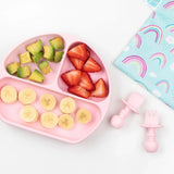 A pink baby plate with fruit sits beside utensils and a Bumkins Reusable Snack Bag, Large 2-Pack: Rainbows & Unicorns on a marble surface.