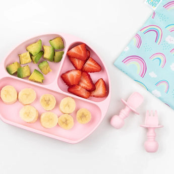 A pink baby plate with fruit sits beside utensils and a Bumkins Reusable Snack Bag, Large 2-Pack: Rainbows & Unicorns on a marble surface.