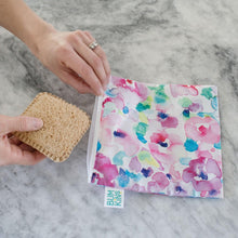 Load image into Gallery viewer, A hand holds a sandwich near a Bumkins Reusable Snack Bag, Large 2-Pack: Watercolor &amp; Brush Strokes on a marble surface.
