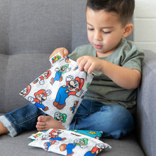 Load image into Gallery viewer, A young child eagerly opens Bumkins Reusable Snack Bag, Large: Super Mario™ &amp; Luigi while sitting on a couch with other similar bags nearby.
