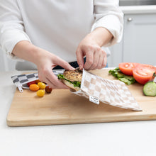 Load image into Gallery viewer, A person places a sandwich into a Roamio by Bumkins Bumkins Reusable Snack Bag, Large 2 Pack: Double Check on a cutting board with vegetables.

