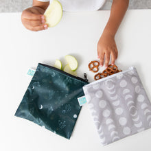 Load image into Gallery viewer, A child's hands hold apple slices as they reach for pretzels from Bumkins Reusable Snack Bag, Large 2 Pack: Desert Night on a white table.
