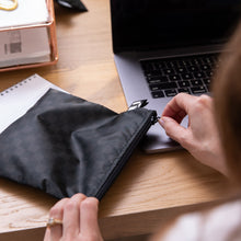 Load image into Gallery viewer, A person unzips a black Roamio by Bumkins Reusable Snack Bag, Large 2 Pack: Rich &amp; Boujee at a desk with work essentials.
