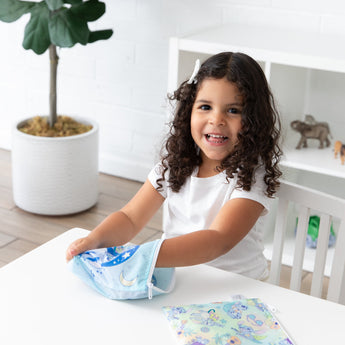 A child with curly hair reaches into a Bumkins Reusable Snack Bag, Large 2 Pack: Stitch Star-ring Up Trouble at a white table.