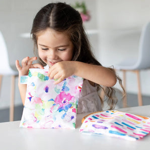 A young girl smiles, holding a Bumkins Reusable Snack Bag, Large 2-Pack: Watercolor & Brush Strokes as the other bag sits on a white table.