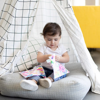 A toddler sits in a checkered fabric tent holding a Bumkins Reusable Snack Bag, Large 2-Pack: Watercolor & Brush Strokes; one sits on their lap.