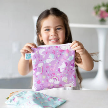 A girl smiles at a white table, holding a Bumkins Reusable Snack Bag, Large 2-Pack: Rainbows & Unicorns; another bag sits in front of her.