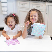 Load image into Gallery viewer, Two smiling children hold Bumkins Reusable Snack Bag, Large 2-Pack: Rainbows &amp; Unicorns at a white table in a kitchen.
