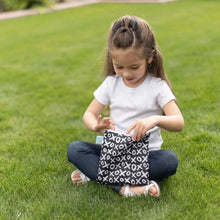 Load image into Gallery viewer, A young girl sitting on grass, wearing a white shirt and holding a Bumkins Reusable Snack Bag, Large 2-Pack: XOXO &amp; Hearts.
