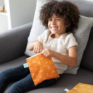 A curly-haired child smiles on a gray couch, holding a Bumkins Reusable Snack Bag, Large 2-Pack: Sunshine and Grounded.