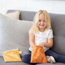 Load image into Gallery viewer, A smiling girl on a gray sofa reaches into a Bumkins Reusable Snack Bag, Large 2-Pack: Grounded, with another bag nearby.
