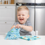 A child smiles at a white table, reaching into a Bumkins Reusable Snack Bag, Large 2-Pack: Ocean Life & Whale Tail with another bag nearby.