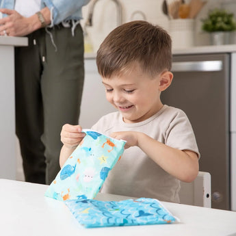 A boy opens Bumkins Reusable Snack Bag, Large 2-Pack: Ocean Life & Whale Tail at a white table, while an adult stands in the background.