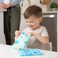 Load image into Gallery viewer, A boy opens Bumkins Reusable Snack Bag, Large 2-Pack: Ocean Life &amp; Whale Tail at a white table, while an adult stands in the background.
