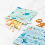Bumkins Reusable Snack Bag, Large 2-Pack: Ocean Life & Whale Tail holds pretzels and veggie sticks on a marble surface.