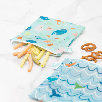 Bumkins Reusable Snack Bag, Large 2-Pack: Ocean Life & Whale Tail holds pretzels and veggie sticks on a marble surface.