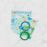 Blue and green teething rings lay on top of Bumkins Reusable Snack Bag, Large 2-Pack: Ocean Life & Whale Tail on a marble background.
