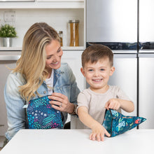Load image into Gallery viewer, A woman and child sit at a white table, smiling with Bumkins Reusable Snack Bag, Large 2-Pack: Jungle &amp; Animal Prints.
