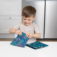 Load image into Gallery viewer, A young boy smiles as he opens Bumkins Reusable Snack Bag, Large 2-Pack: Jungle &amp; Animal Prints at a white table in a kitchen.
