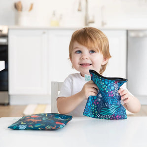 A toddler smiles while holding a Bumkins Reusable Snack Bag, Large 2-Pack: Jungle & Animal Prints with another bag on a white table.