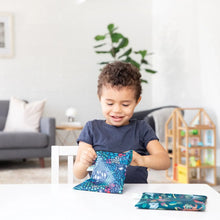 Load image into Gallery viewer, A child beams while opening a Bumkins Reusable Snack Bag, Large 2-Pack: Jungle &amp; Animal Prints at a white table in a cozy living room.
